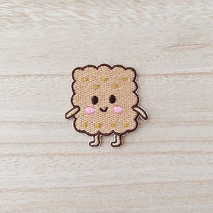 May include: A brown embroidered patch of a smiling cartoon biscuit with a happy face and pink cheeks.