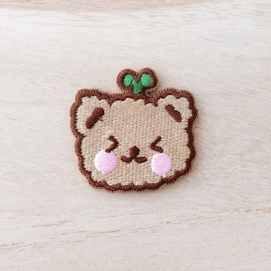 May include: Embroidered brown bear patch with pink cheeks and a green leaf on top.
