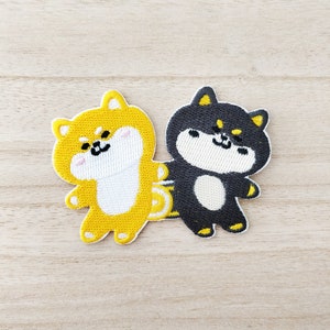 May include: Two embroidered iron-on patches of cartoon Shiba Inu dogs. One is yellow and the other is black. Both have white faces and are smiling.