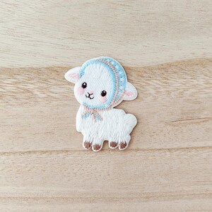 May include: Embroidered patch of a white lamb wearing a blue bonnet with a pink trim and a matching scarf. The lamb has brown hooves and a sweet expression. The patch is on a light wood grain surface.