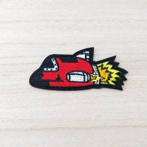 May include: Red and black embroidered iron-on patch of a cartoon spaceship with yellow flames coming out of the back.
