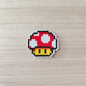 May include: A pixelated patch featuring a red mushroom with white spots on the cap, a yellow stem, and black eyes. The patch has a white border and is likely an iron-on or sew-on applique. The design is reminiscent of classic video games.