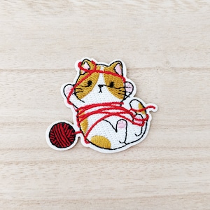 May include: Embroidered iron-on patch of a white cat with orange and yellow markings tangled in red yarn with a red ball of yarn next to it.