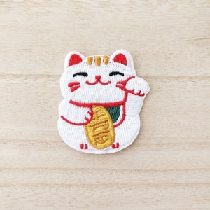 Peut inclure: Patch brodé blanc représentant un Maneki-Neko japonais souriant, ou chat porte-bonheur, avec une patte levée et tenant une pièce d'or. Le chat a les joues rouges et un collier rouge.