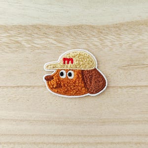 May include: Embroidered patch of a cartoon dog wearing a beige baseball cap with a red "M". The dog has brown fur, a brown ear, and large black eyes. The patch has a white border.