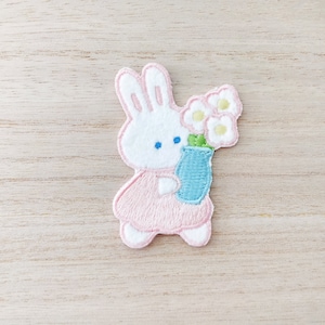 Peut inclure: Un lapin blanc brodé tenant un vase bleu avec trois fleurs blanches. Le lapin porte une robe rose.