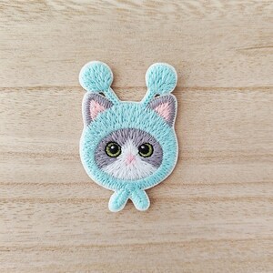 May include: Embroidered patch of a cartoon cat wearing a light blue hat with pom-poms. The cat has green eyes, a pink nose, and grey and white fur. The patch is on a light brown wooden surface.