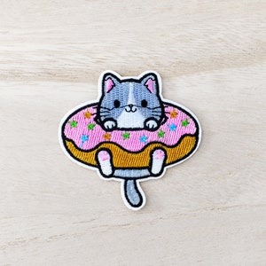 Peut inclure: Patch brodée thermocollante représentant un chat gris assis dans un donut rose avec des pépites colorées.