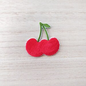 May include: A red embroidered cherry patch with green stems and leaves. The patch is ideal for adding a touch of whimsy to clothing, bags, or other crafts.