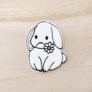 May include: White embroidered iron-on patch of a bunny holding a flower. The bunny is sitting with its legs crossed and has a pink nose and black eyes.