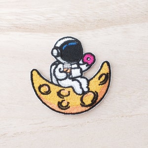 May include: An embroidered patch of an astronaut sitting on a yellow crescent moon, holding a doughnut in one hand and a cup in the other.