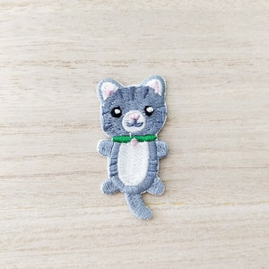 May include: An iron-on patch featuring an embroidered grey cat wearing a green collar. The cat has white fur on its chest and belly.