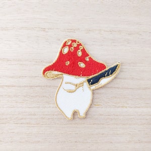 Peut inclure: Écusson brodé représentant un champignon de dessin animé avec un chapeau rouge à pois blancs, un corps blanc et un contour doré. Le champignon tient un couteau bleu et or. Le patch est conçu pour être appliqué sur des vêtements ou des accessoires.