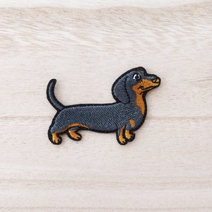 Peut inclure: Patch brodé représentant un teckel noir et fauve avec un nez brun et de grands yeux. Le teckel est tourné vers la droite et a la langue qui pend.