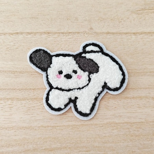 May include: A white and black dog-shaped patch with a light blue border. The dog has black ears and spots, pink cheeks, and a fluffy texture. The patch is on a light brown wooden surface.