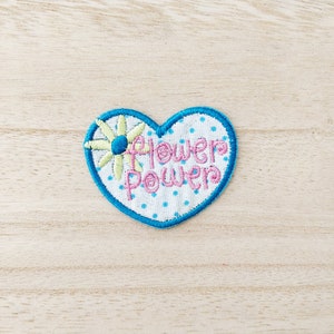 May include: A blue heart-shaped patch with white polka dots and the text "Flower Power" in pink and yellow embroidery. A yellow flower with a blue centre is in the top left corner.