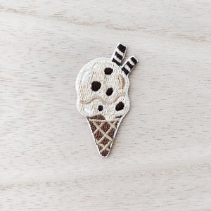 Peut inclure: Un patch brodé représentant un cornet de glace brun et blanc avec des pépites de chocolat et un topping rayé brun et blanc.
