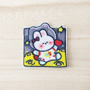 Peut inclure: Patch brodée thermocollante représentant un lapin blanc portant une combinaison d'astronaute avec un cœur rouge sur la poitrine. Le lapin est assis sur une lune jaune avec un fond étoilé bleu et gris.