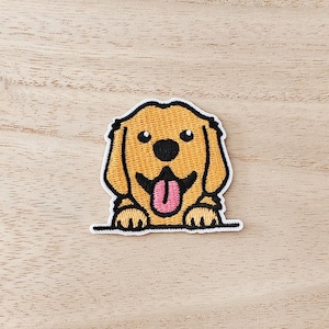 May include: Embroidered patch of a smiling golden retriever dog with a pink tongue. The patch has a white border and is set against a light wood grain background. The dog's fur is a light brown colour.