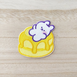 May include: Embroidered patch depicting a yellow pancake with syrup and a white cloud-like character with purple detailing. The patch is set against a light wood grain background.