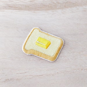 May include: A yellow embroidered patch depicting a slice of bread with a pat of butter on top. The patch is edged with a white border and a yellow outline.