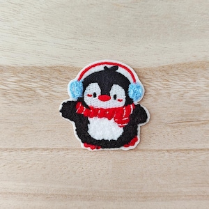 May include: Embroidered penguin patch featuring a red scarf, blue earmuffs, and a red and white headband. The penguin has a white belly, black wings, and red feet. Ideal for adding a touch of winter charm to clothing or accessories.