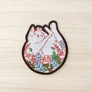 May include: Embroidered patch featuring a white cat in a relaxed pose, surrounded by pink and blue flowers and green leaves. The patch has a black border and is set against a light wood grain background.