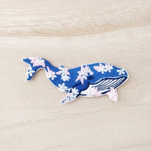 Peut inclure: Un patch brodé représentant une baleine bleue avec des fleurs blanches et roses. La baleine est tournée vers la droite et a un contour blanc.