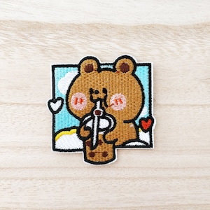 Peut inclure: Patch brodé représentant un ours brun aux joues roses buvant un thé à la bulle. L'ours est entouré de nuages blancs et de cœurs sur un fond bleu.