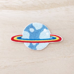 May include: A blue and white embroidered patch of a planet with a red, yellow, and blue ring around it.