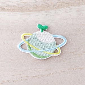 May include: Green and white embroidered planet with a blue and yellow ring around it. The planet has a small green sprout on top.