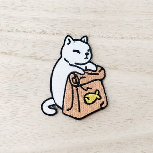 May include: White embroidered cat patch with a brown bag featuring a yellow fish design. The cat is holding the bag with its paws.