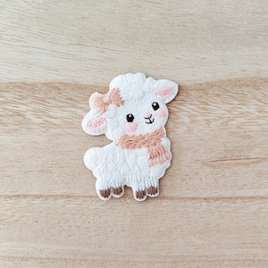 May include: A white embroidered lamb patch with a pink bow, scarf, and blush. The lamb has brown hooves and a smiling face. The patch is on a light brown wooden surface.