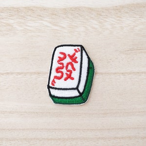 May include: Embroidered Mahjong tile patch. The patch has red Chinese characters on a white background, with a black and green border. The rectangular patch with rounded edges is designed for application to fabric.