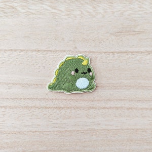 May include: Green embroidered iron-on patch of a cartoon dinosaur with a yellow horn, white belly, and pink cheeks.