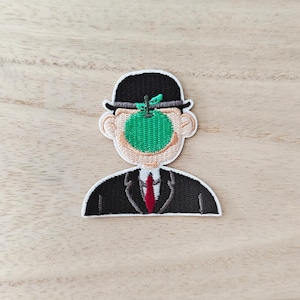 Peut inclure: Écusson brodé représentant un homme en chapeau melon noir et costume, avec une pomme verte cachant son visage. Le patch a une bordure crème et une cravate rouge. Le design est inspiré de l'œuvre de René Magritte.
