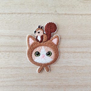 May include: Embroidered patch featuring a brown cat with a squirrel on its head. The cat has green eyes, pink accents, and a white muzzle. The squirrel is brown with a fluffy tail. The patch is on a light wood grain surface.