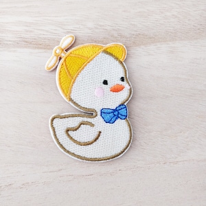 May include: Embroidered patch of a cartoon duckling wearing a yellow aviator cap and a blue bow tie. The duckling has a white body, pink cheeks, and an orange beak. The patch is outlined in brown and is on a light wood surface.