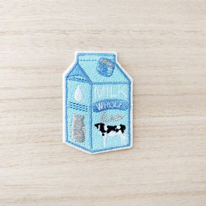 Peut inclure: Écusson brodé en forme de carton de lait bleu clair. Le patch présente le mot "MILK" et une illustration de vache. Des détails supplémentaires incluent une goutte de lait et un petit récipient.