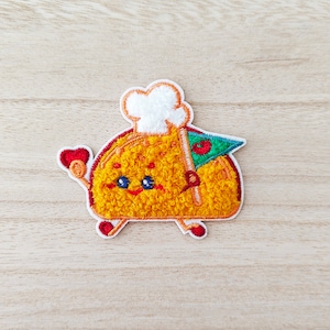May include: A yellow embroidered patch of a cartoon taco wearing a white chef's hat and holding a green flag with a red cherry on it.