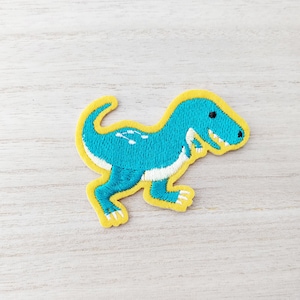 May include: A turquoise blue embroidered dinosaur patch with yellow outline. The dinosaur is walking with its mouth open and has white teeth.