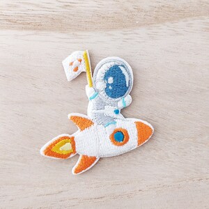 May include: An embroidered iron-on patch featuring a white rocket with orange flames and an astronaut in a white spacesuit holding a flag.