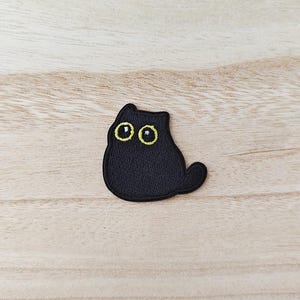 May include: A black cat patch with yellow eyes and a small tail. The patch is embroidered and has a simple, cartoon-like design. The patch is on a light wood grain surface.