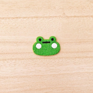 May include: A green embroidered frog patch with white accents. The frog has white eyes, black pupils, and a black horizontal line for a mouth. The patch is on a light brown wooden surface.