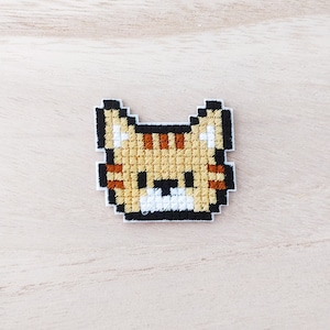 May include: A pixelated embroidered patch of a cat with orange and white fur. The cat has black eyes and a black nose.