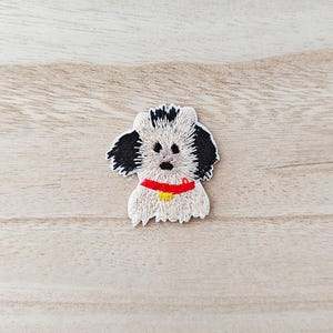 May include: Embroidered dog patch with a white body, black ears, and a red collar. The dog has a yellow tag on its collar. The patch is on a light brown wooden surface.