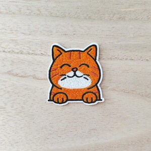 Peut inclure: Écusson brodé représentant un chat tabby orange souriant avec des rayures noires. Le chat a un ventre et des pattes blancs, et est bordé de noir. L'écusson est sur une surface en bois clair.
