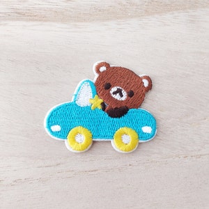 May include: Embroidered iron-on patch featuring a brown bear driving a blue car with yellow wheels.
