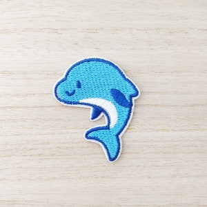 May include: A cheerful, embroidered patch featuring a light blue dolphin with a white belly and dark blue accents. The dolphin has a smiling face and is outlined in white. The patch is designed for application on clothing or accessories.