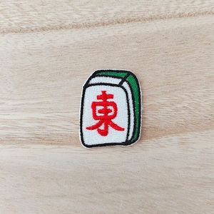 May include: A rectangular embroidered patch shaped like a mahjong tile. The patch features a white background with a red Japanese kanji character for east, bordered by a thick black outline and a green accent stripe along the top and right side.